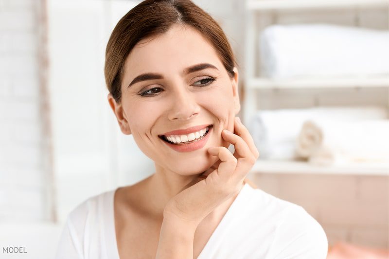 Middle aged woman in white shirt, smiling and resting her hand on her jaw.